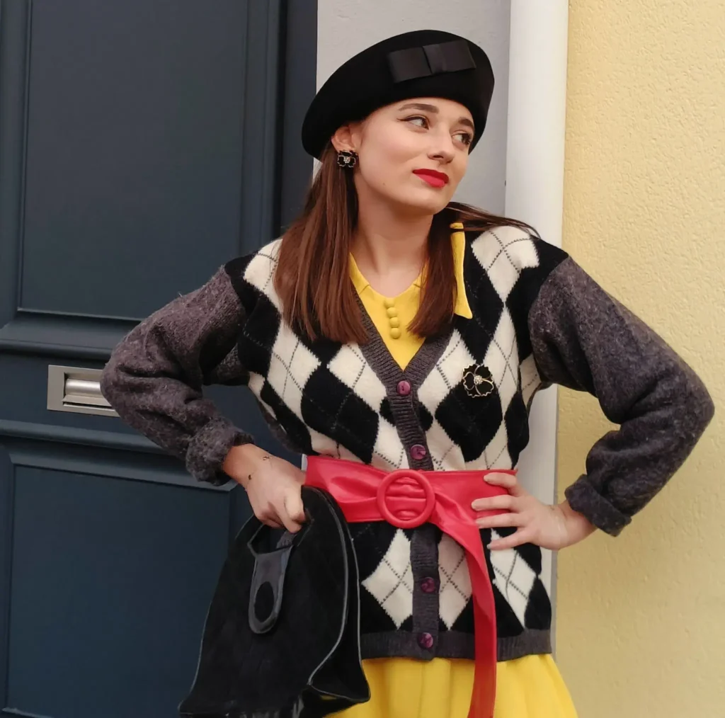 Femme adossée à un mur, les mains sur les hanches, habillée chic avec un chapeau noir vintage.