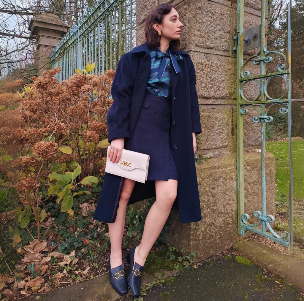 Femme vintage au style années 50, vêtue de bleu et d'une pochette blanc cassé à la main, vue de face.